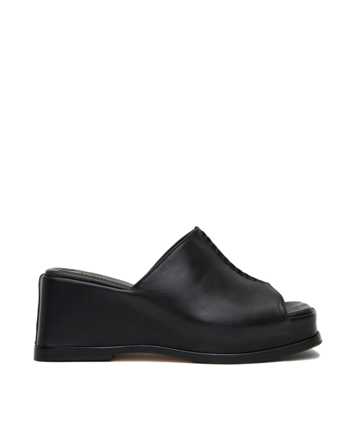 Women's wedge platform mule slide-on shoes made from buttery soft premium European black leather. The shoe design features a wholecut upper, meaning that it's been cut in one continuous piece. The toe and heel of the shoe are a chiseled square shape. The insole is fully padded for all-day comfort and the nubuck sole features non-slip rubber inserts. The perfect 90s inspired cool girl shoe for spring summer and fall.