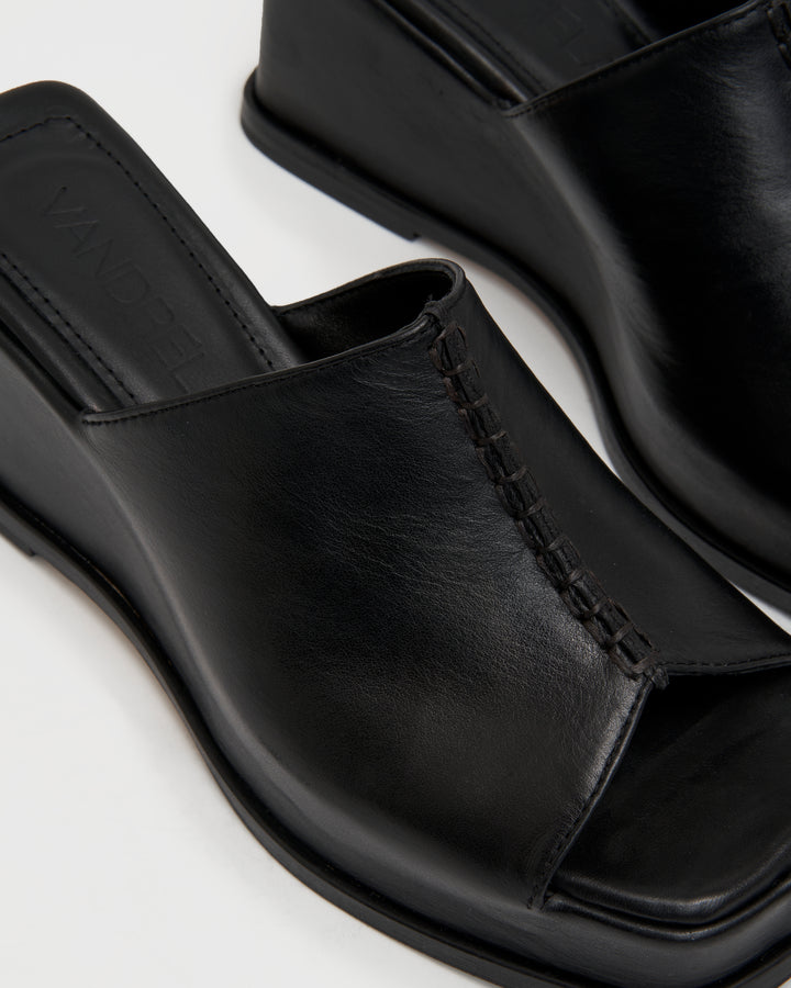 Women's wedge platform mule slide-on shoes made from buttery soft premium European black leather. The shoe design features a wholecut upper, meaning that it's been cut in one continuous piece. The toe and heel of the shoe are a chiseled square shape. The insole is fully padded for all-day comfort and the nubuck sole features non-slip rubber inserts. The perfect 90s inspired cool girl shoe for spring summer and fall.