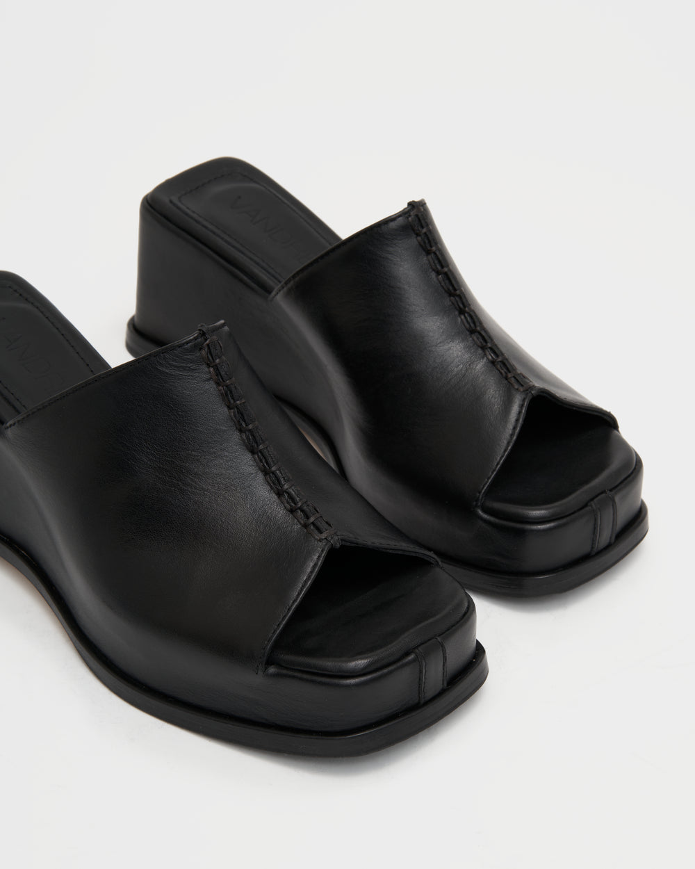 Women's wedge platform mule slide-on shoes made from buttery soft premium European black leather. The shoe design features a wholecut upper, meaning that it's been cut in one continuous piece. The toe and heel of the shoe are a chiseled square shape. The insole is fully padded for all-day comfort and the nubuck sole features non-slip rubber inserts. The perfect 90s inspired cool girl shoe for spring summer and fall.