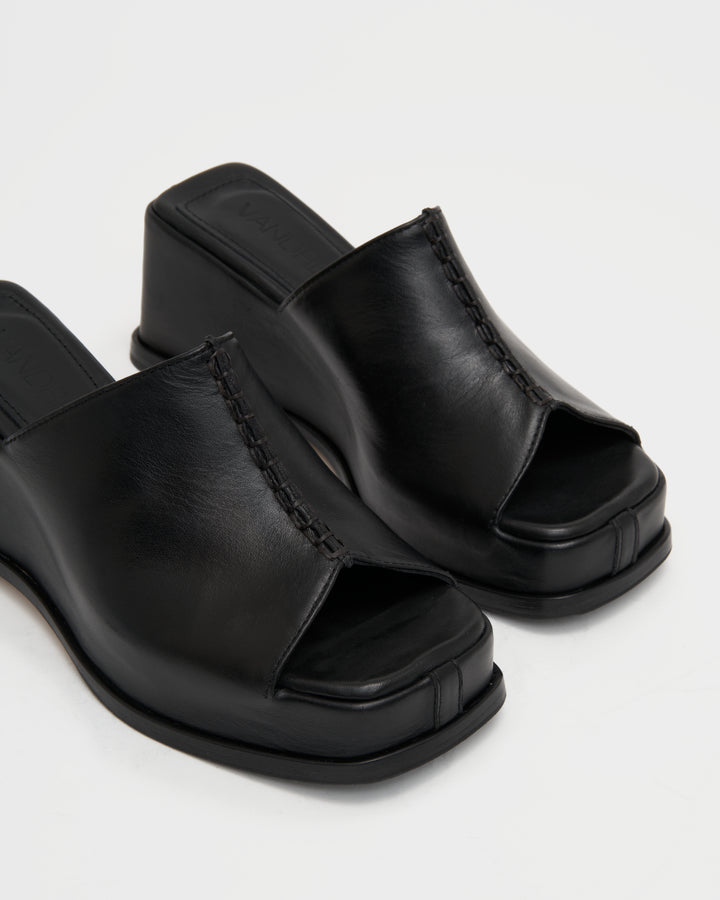 Women's wedge platform mule slide-on shoes made from buttery soft premium European black leather. The shoe design features a wholecut upper, meaning that it's been cut in one continuous piece. The toe and heel of the shoe are a chiseled square shape. The insole is fully padded for all-day comfort and the nubuck sole features non-slip rubber inserts. The perfect 90s inspired cool girl shoe for spring summer and fall.